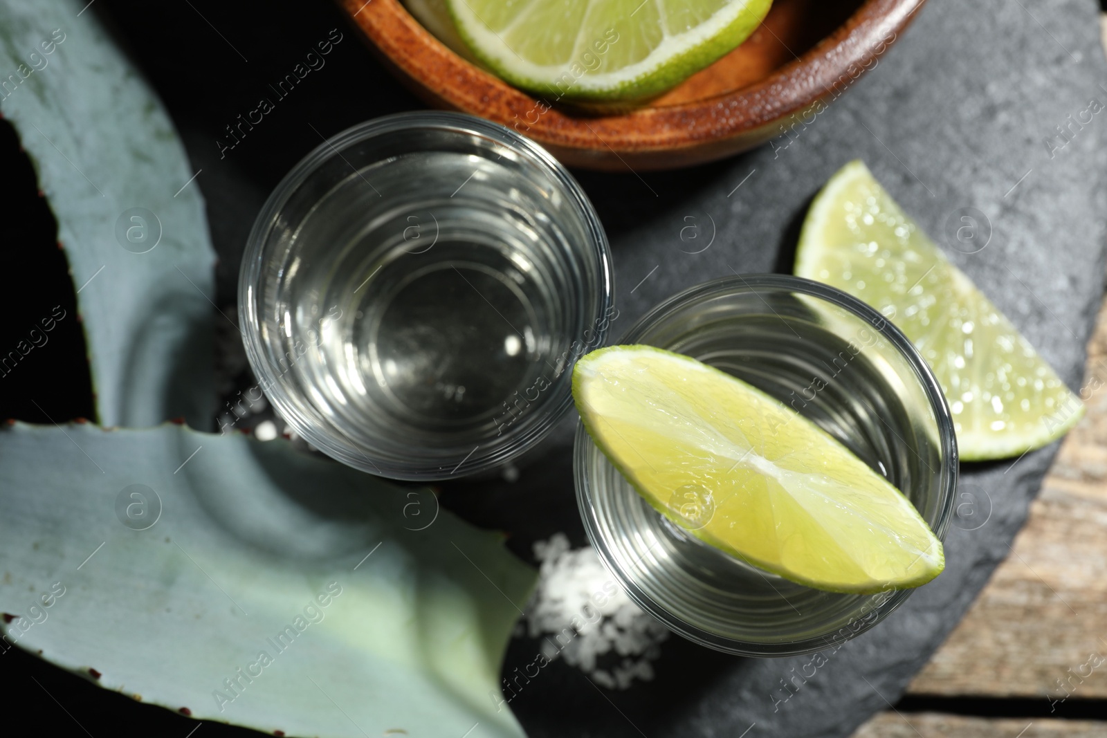 Photo of Tequila shots with lime slices, salt and agave leaves on wooden table, flat lay