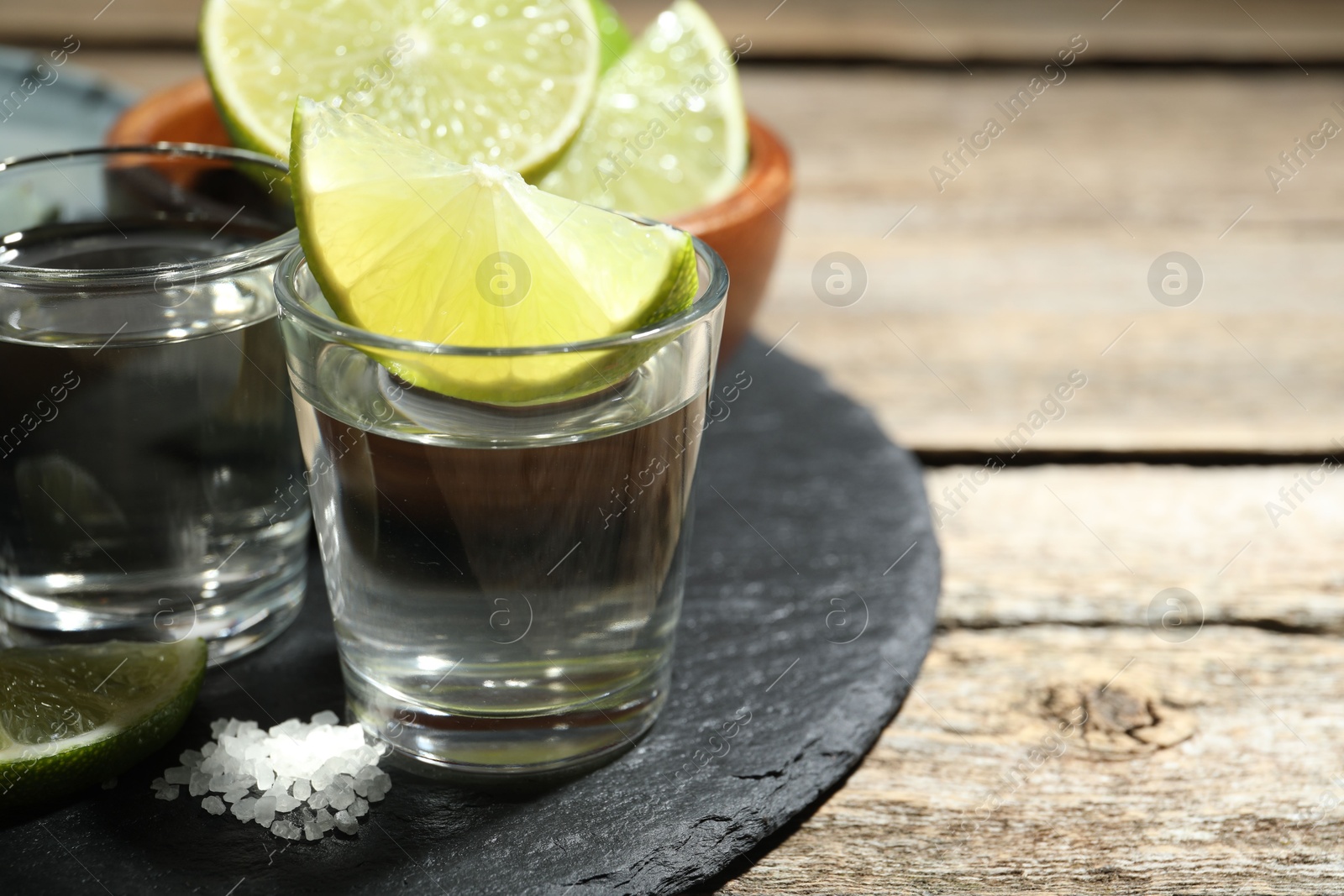 Photo of Tequila shots with lime slices and salt on wooden table, closeup. Space for text