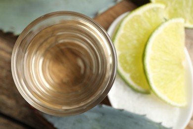 Photo of Tequila shot with lime slices, salt and agave leaves on wooden table, flat lay