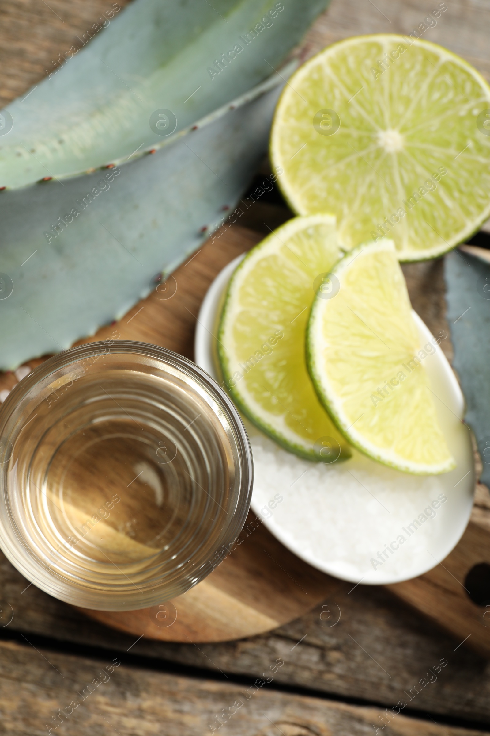 Photo of Tequila shot with lime slices, salt and agave leaves on wooden table, flat lay