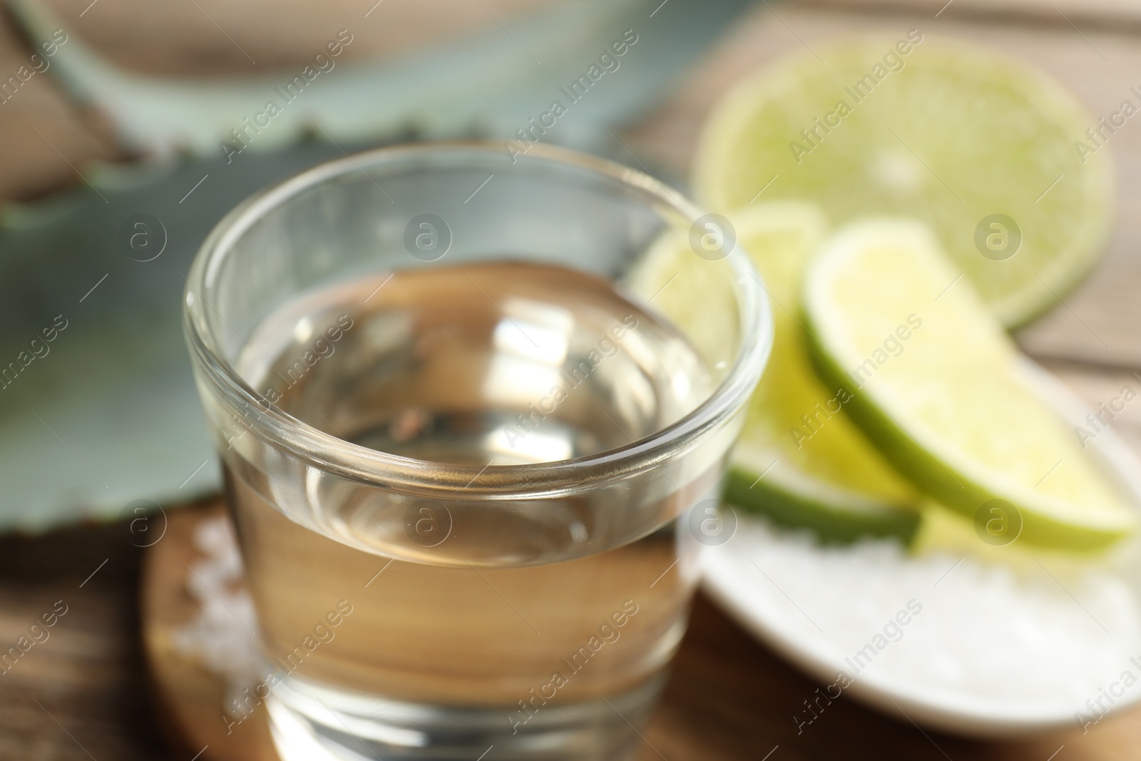 Photo of Tequila shot with lime slices and agave leaves on wooden table, closeup