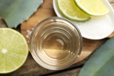 Photo of Tequila shot with lime slices, salt and agave leaves on wooden table, flat lay