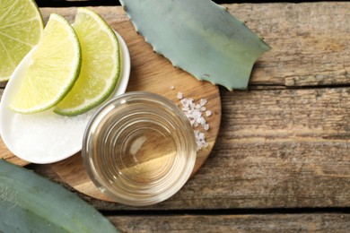 Photo of Tequila shot with lime slices, salt and agave leaves on wooden table, flat lay. Space for text