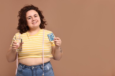 Photo of Happy plus size woman holding blister of weight loss supplements and glass of water on beige background. Space for text