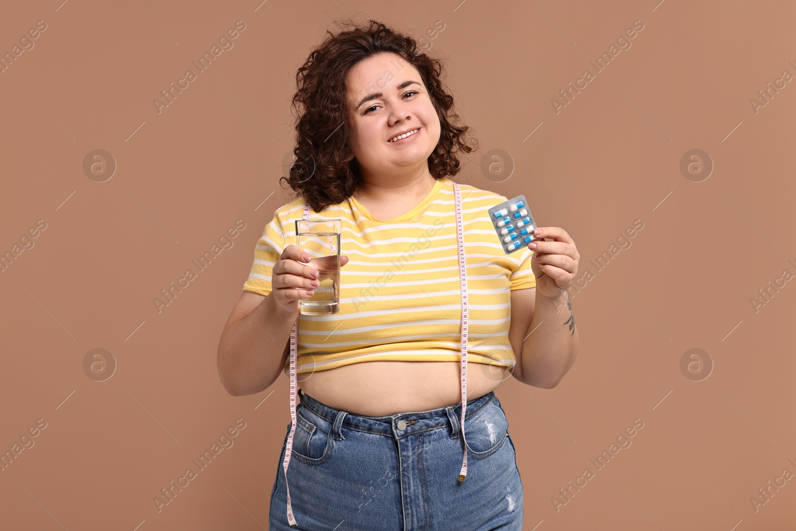 Photo of Happy plus size woman holding blister of weight loss supplements and glass of water on beige background