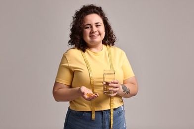 Photo of Happy plus size woman with weight loss supplements and glass of water on grey background