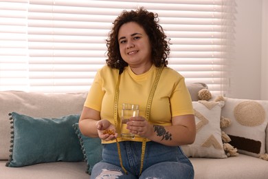 Photo of Happy plus size woman with pile of weight loss supplements and glass of water on sofa at home