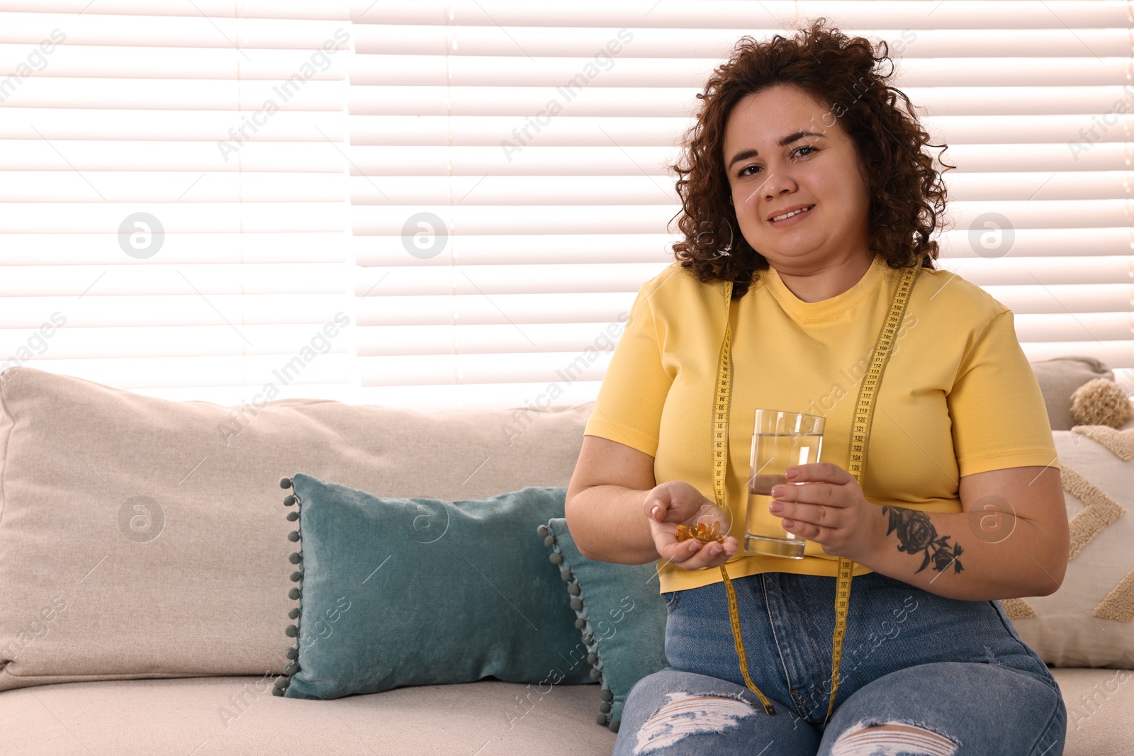 Photo of Happy plus size woman with pile of weight loss supplements and glass of water on sofa at home. Space for text