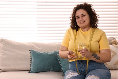 Photo of Happy plus size woman with pile of weight loss supplements and glass of water on sofa at home. Space for text