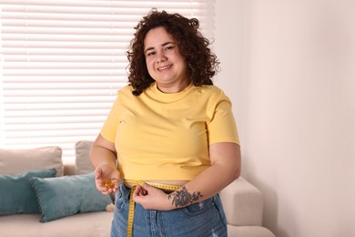 Photo of Happy plus size woman measuring waist with tape and holding pile of weight loss supplements at home