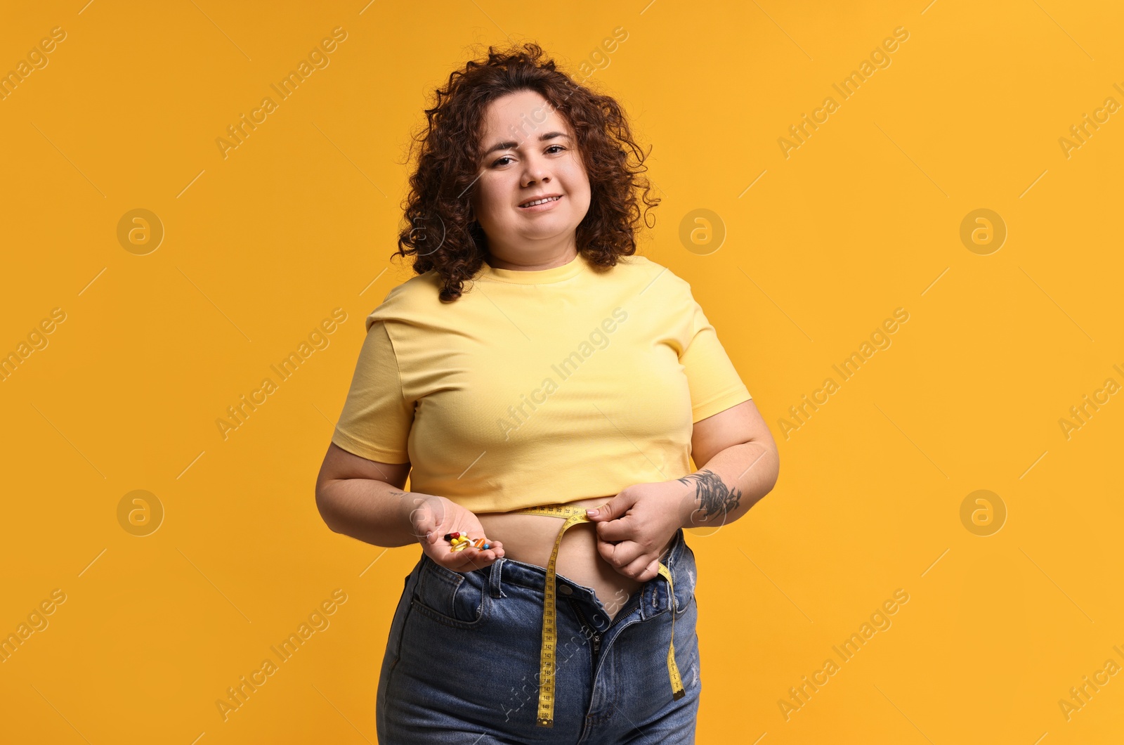 Photo of Happy plus size woman measuring waist with tape and holding pile of weight loss supplements on orange background