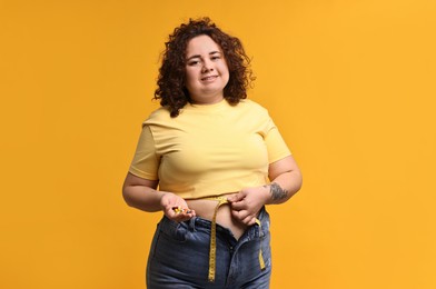 Photo of Happy plus size woman measuring waist with tape and holding pile of weight loss supplements on orange background