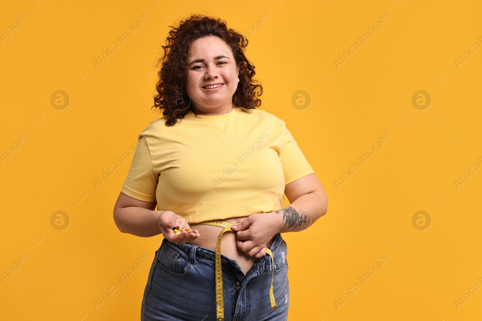 Photo of Happy plus size woman measuring waist with tape and holding pile of weight loss supplements on orange background