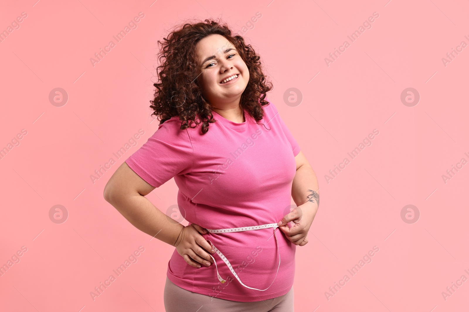 Photo of Weight loss. Happy plus size woman measuring waist with tape on pink background