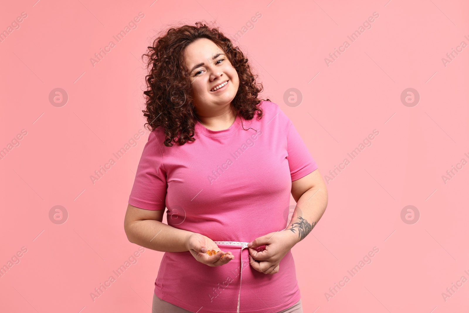 Photo of Happy plus size woman measuring waist with tape and holding pile of weight loss supplements on pink background