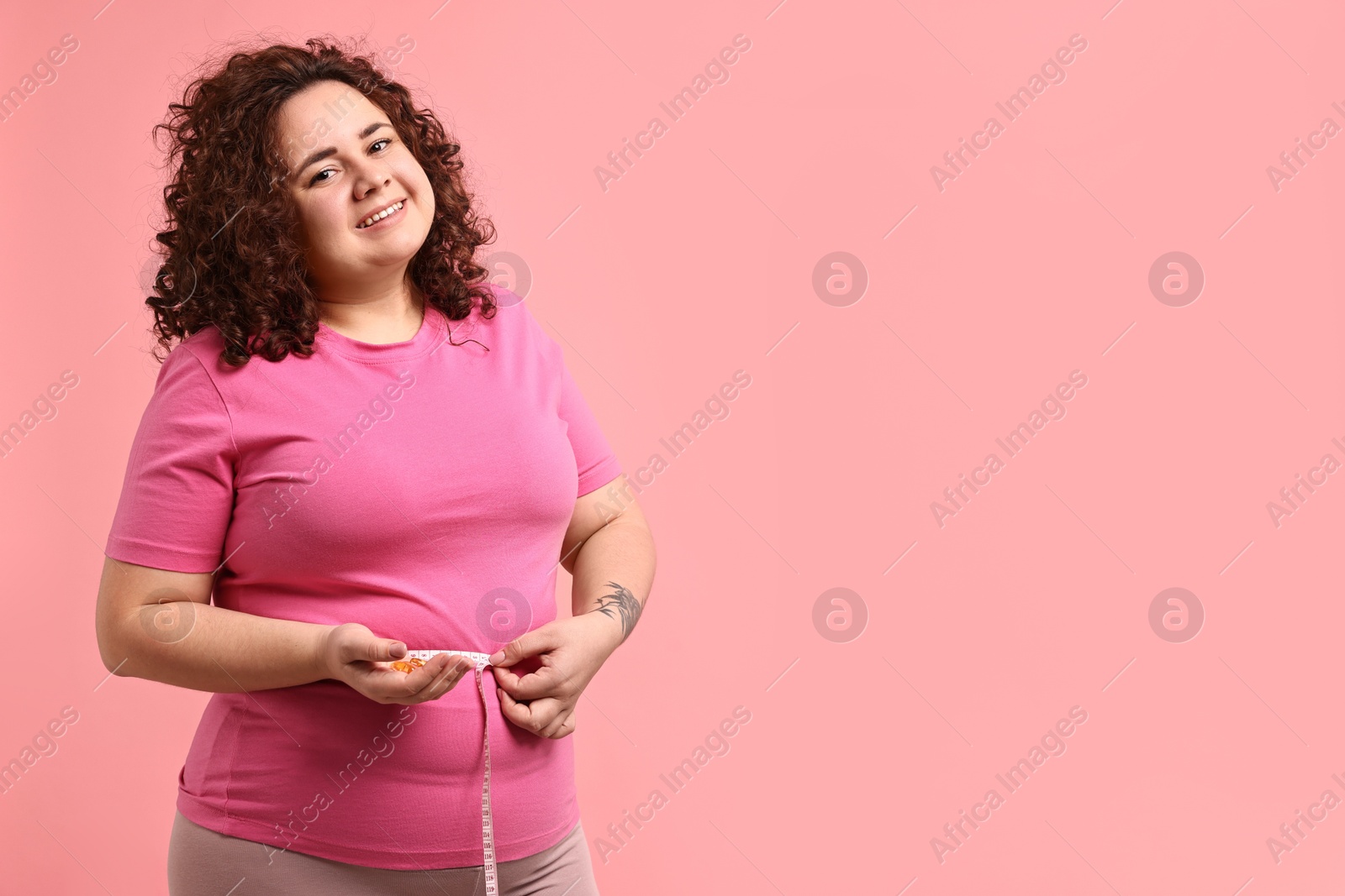 Photo of Happy plus size woman measuring waist with tape and holding pile of weight loss supplements on pink background. Space for text