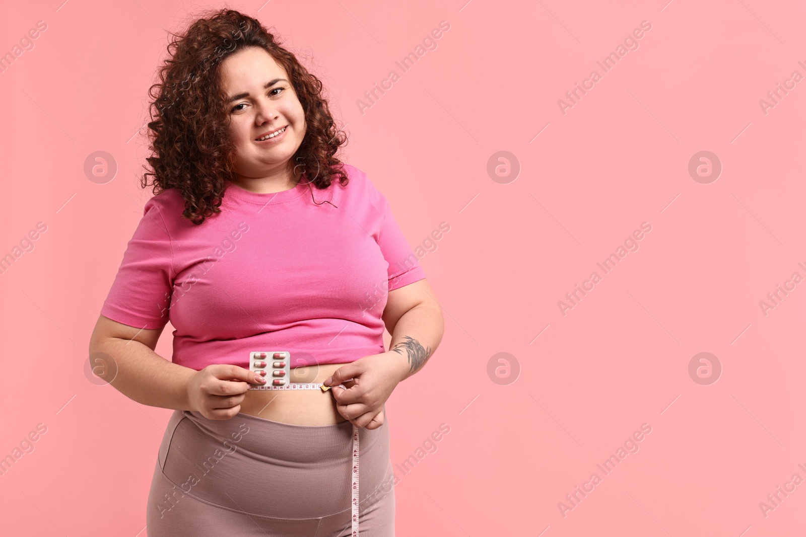 Photo of Happy plus size woman measuring waist with tape and holding blister of weight loss supplements on pink background. Space for text