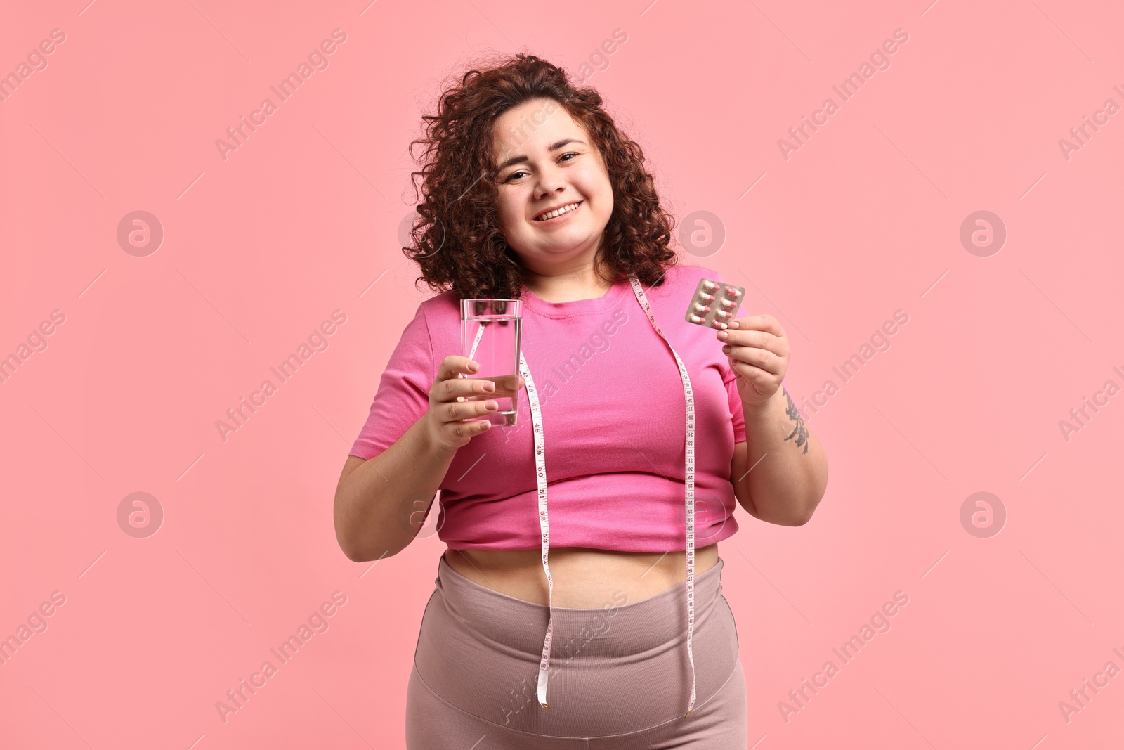 Photo of Happy plus size woman holding blister of weight loss supplements and glass of water on pink background