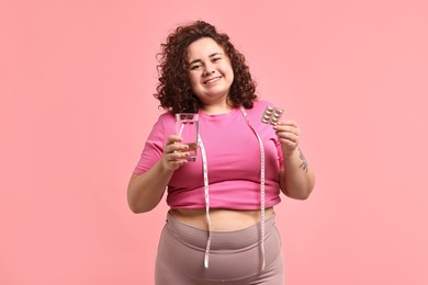 Photo of Happy plus size woman holding blister of weight loss supplements and glass of water on pink background