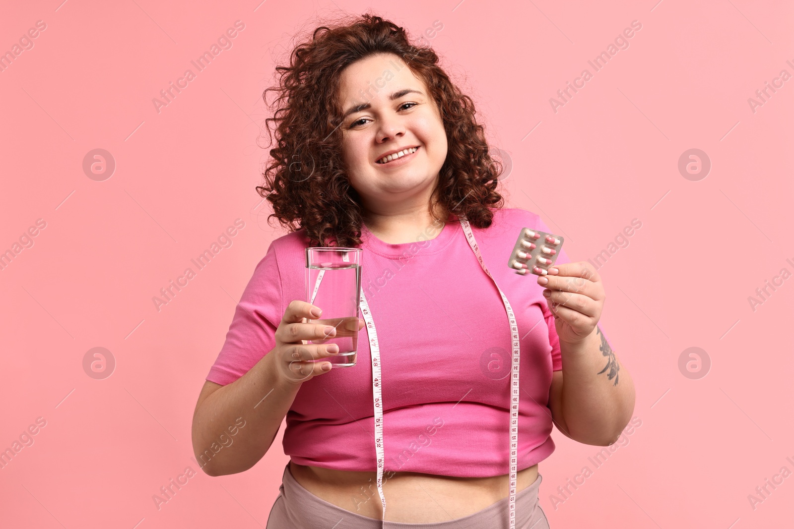 Photo of Happy plus size woman holding blister of weight loss supplements and glass of water on pink background