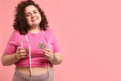 Photo of Happy plus size woman holding blister of weight loss supplements and glass of water on pink background. Space for text