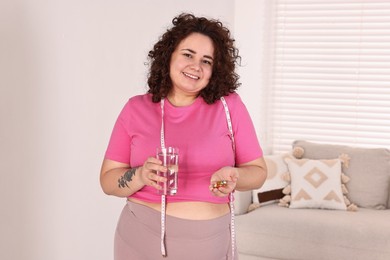 Photo of Happy plus size woman holding pile of weight loss supplements and glass with water at home