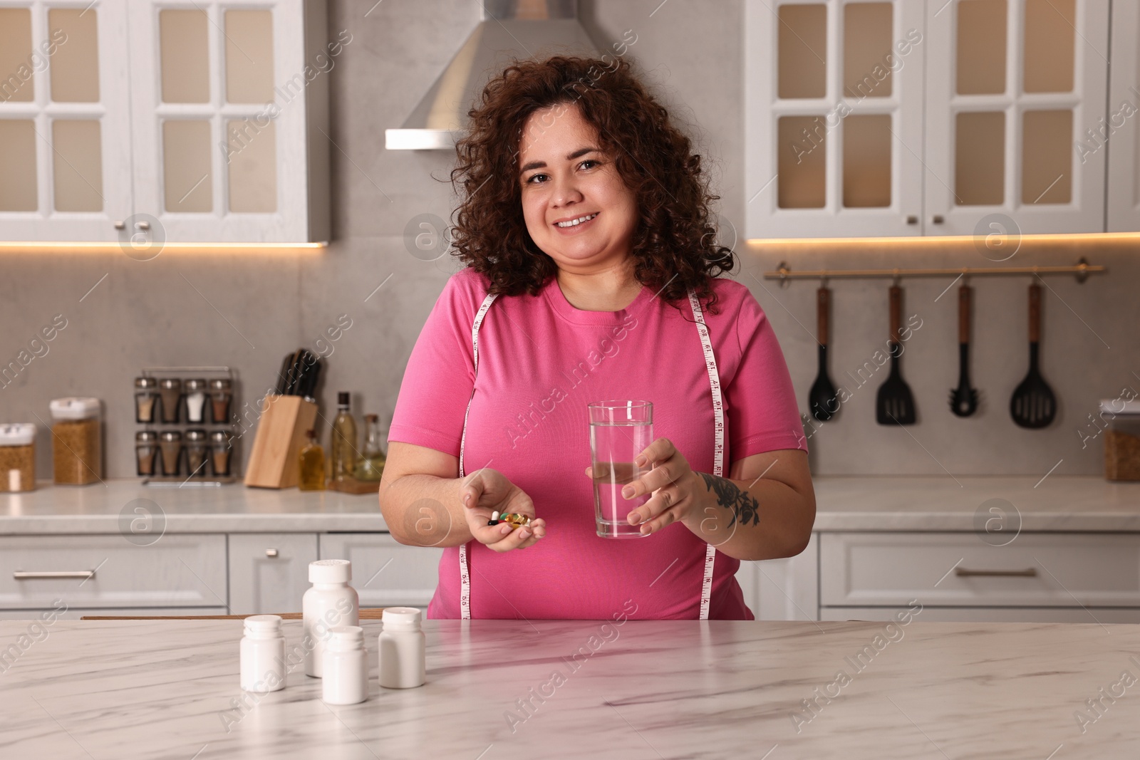 Photo of Happy plus size woman with weight loss supplements and glass of water at marble countertop in kitchen