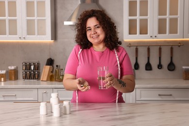 Photo of Happy plus size woman with weight loss supplements and glass of water at marble countertop in kitchen