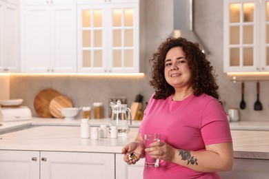 Photo of Happy plus size woman holding pile of weight loss supplements and glass with water in kitchen. Space for text