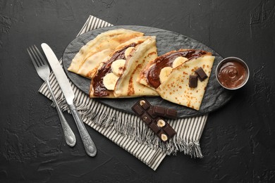 Tasty crepes with chocolate butter, banana and cutlery on black table, flat lay