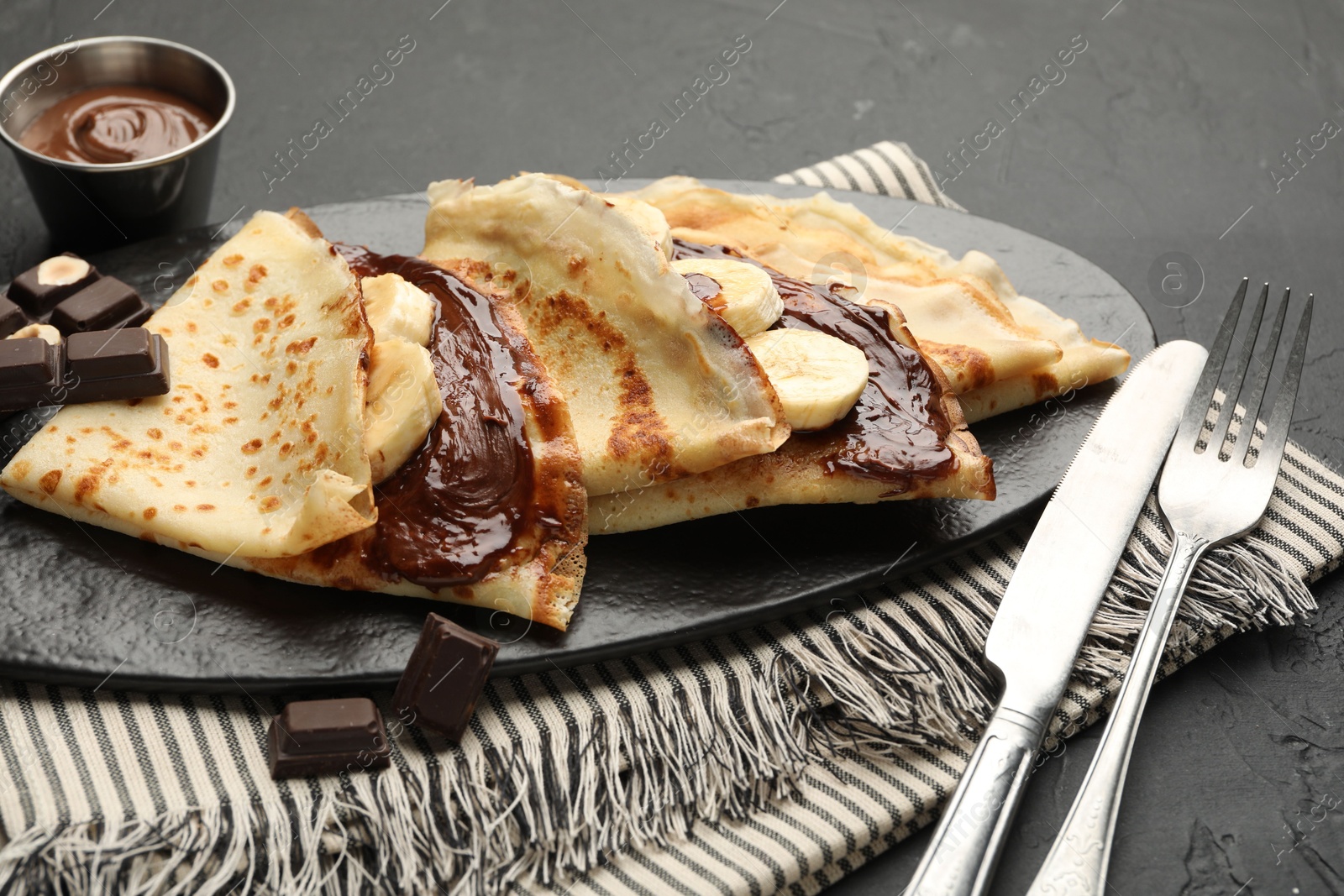 Photo of Tasty crepes with chocolate butter, banana and cutlery on black table, closeup