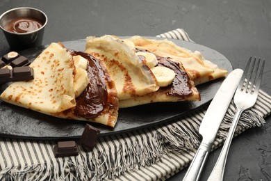 Photo of Tasty crepes with chocolate butter, banana and cutlery on black table, closeup