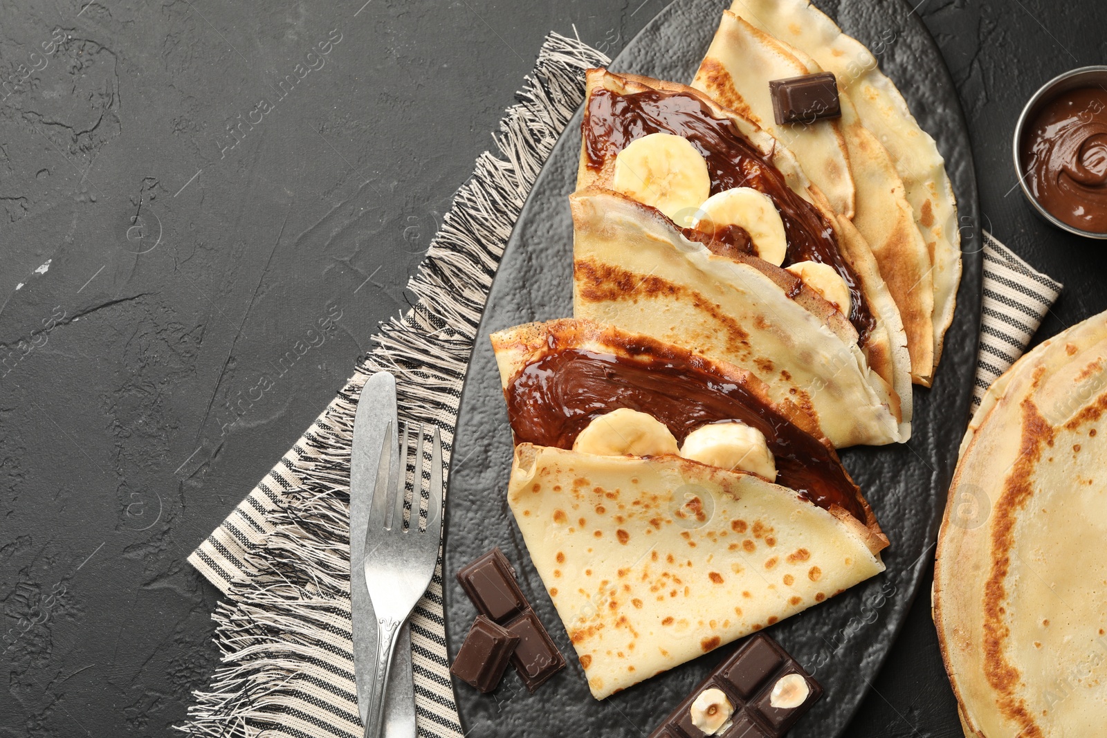 Photo of Tasty crepes with chocolate butter, banana and cutlery on black table, flat lay. Space for text