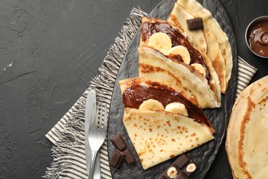 Photo of Tasty crepes with chocolate butter, banana and cutlery on black table, flat lay. Space for text