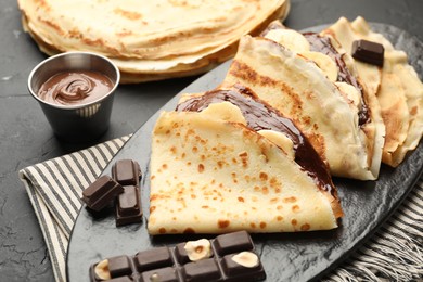 Photo of Tasty crepes with chocolate butter and banana on black table, closeup