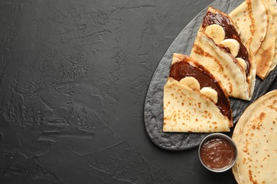 Photo of Tasty crepes with chocolate butter and banana on black table, flat lay. Space for text