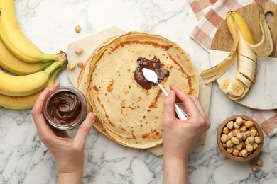 Woman spreading tasty chocolate butter onto crepe at white marble table, top view