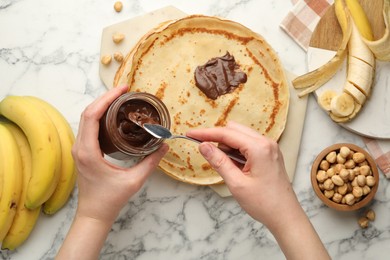 Photo of Woman spreading tasty chocolate butter onto crepe at white marble table, top view