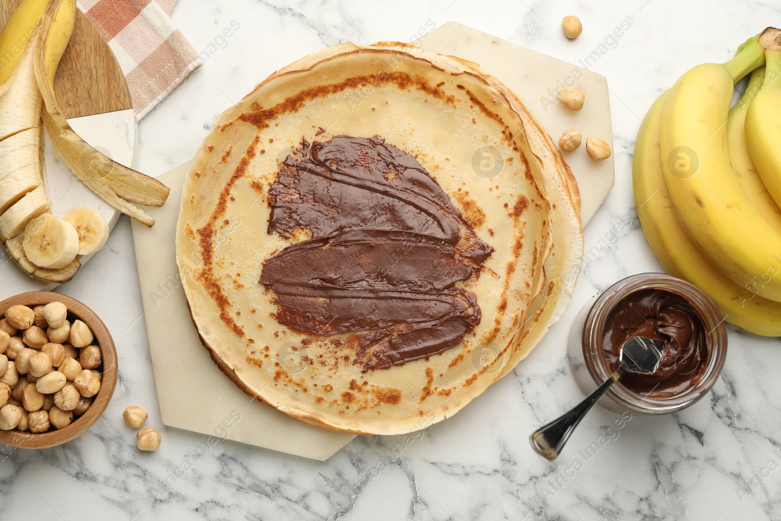 Photo of Tasty crepes with chocolate butter, bananas and hazelnuts on white marble table, flat lay