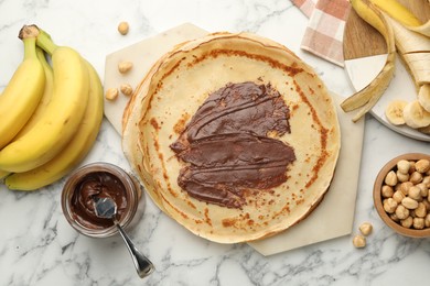 Tasty crepes with chocolate butter, bananas and hazelnuts on white marble table, flat lay