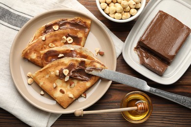 Photo of Tasty crepes with chocolate butter, nuts, honey and knife on wooden table, flat lay