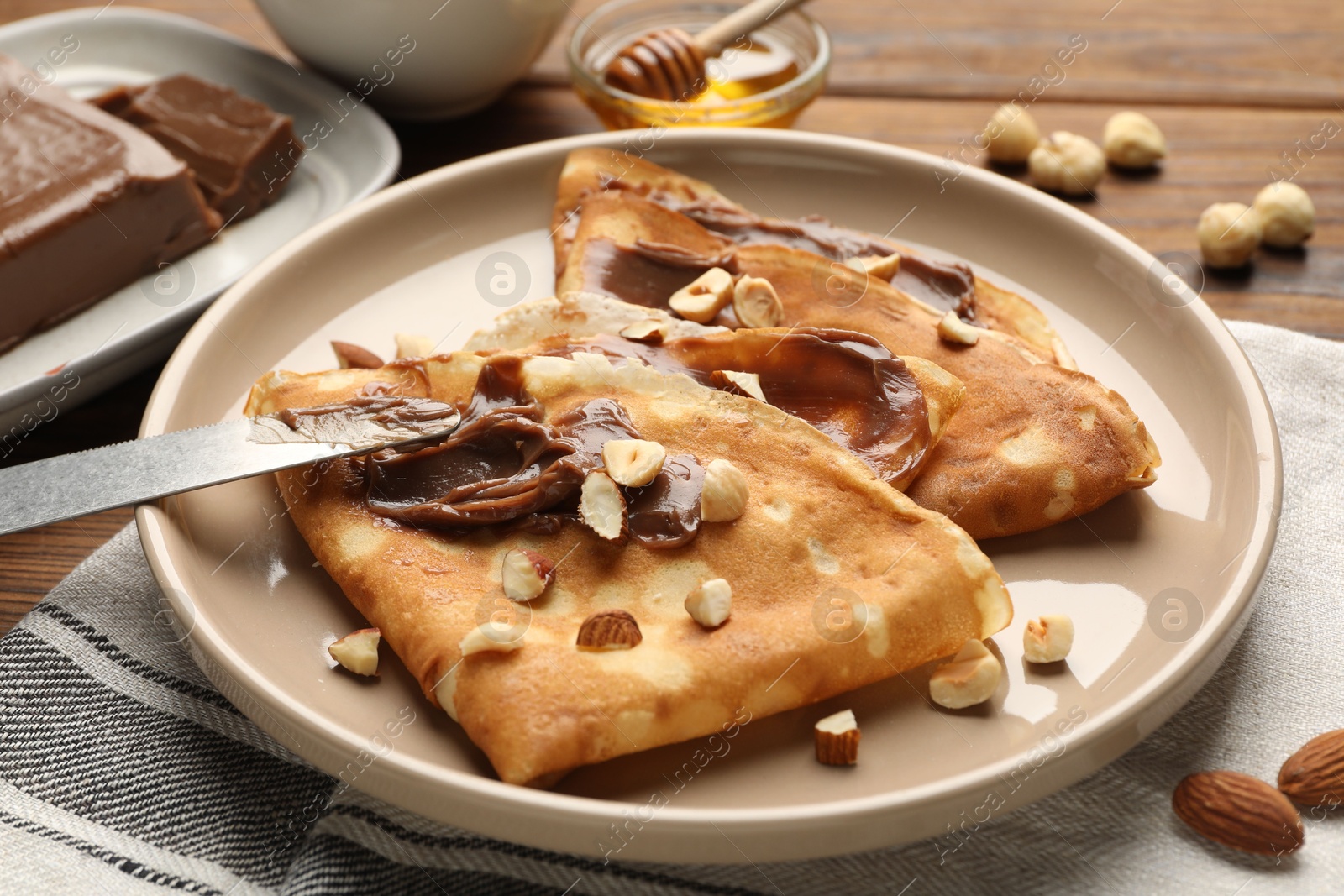 Photo of Tasty crepes with chocolate butter, nuts, honey and knife on wooden table, closeup