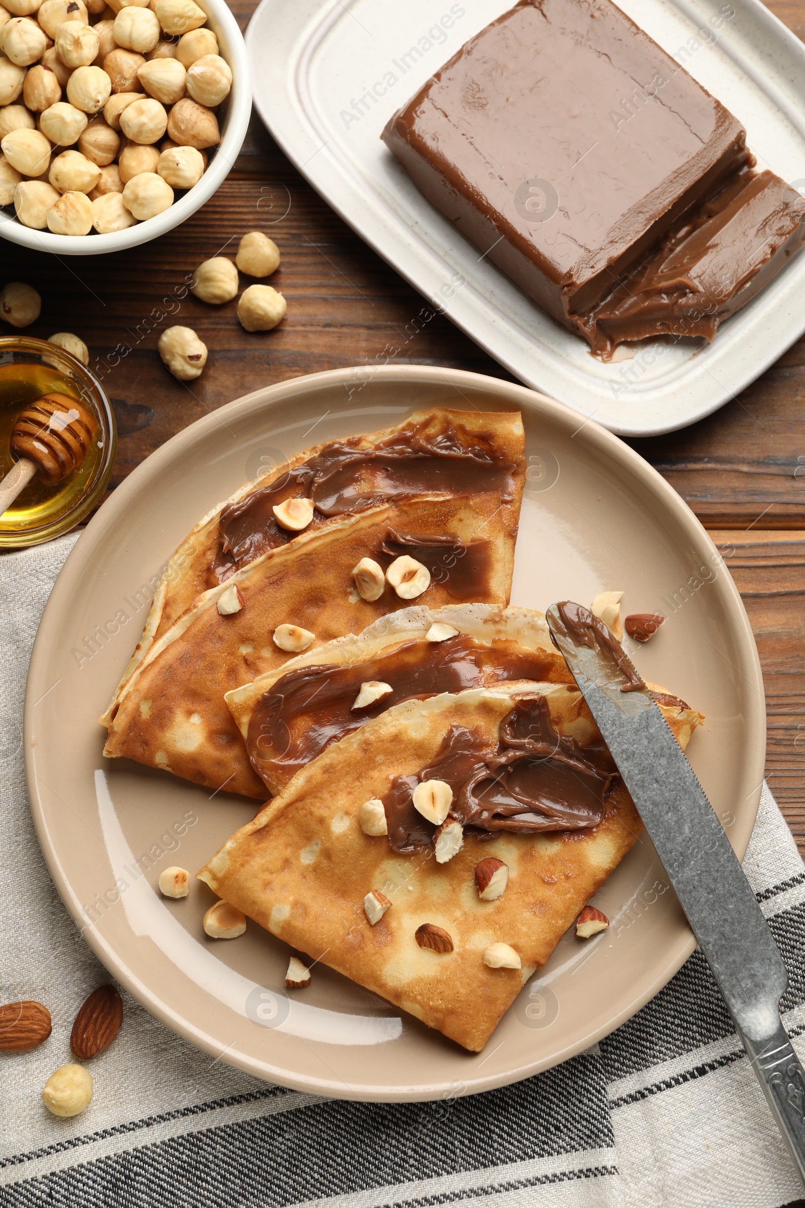 Photo of Tasty crepes with chocolate butter, nuts, honey and knife on wooden table, flat lay