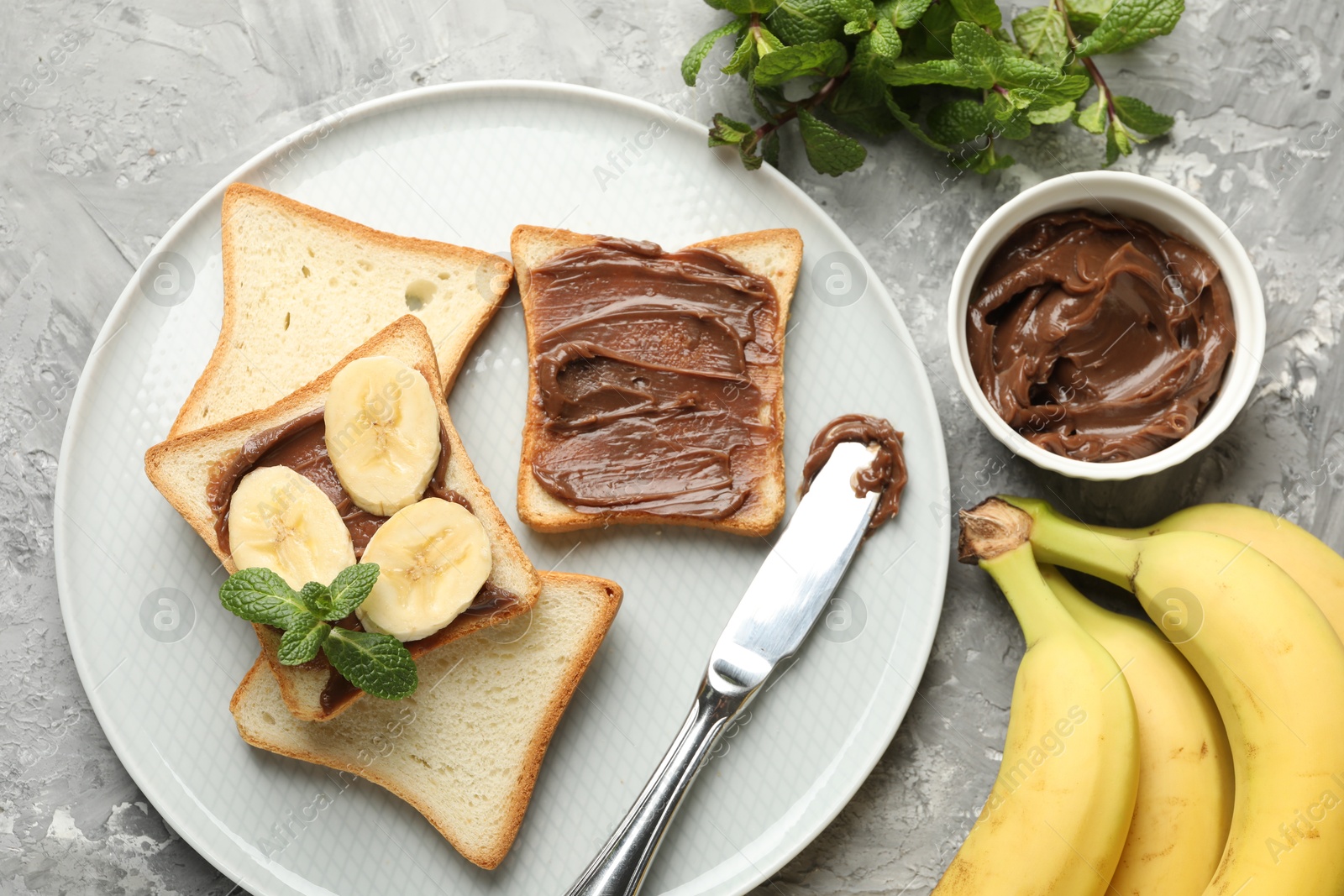 Photo of Sandwiches with chocolate butter, bananas, mint and knife on grey table, flat lay