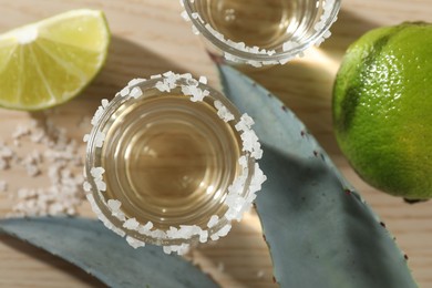 Photo of Tequila shots with salt, limes and agave leaves on wooden table, flat lay