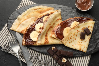 Photo of Tasty crepes with chocolate butter, banana and cutlery on black table, flat lay