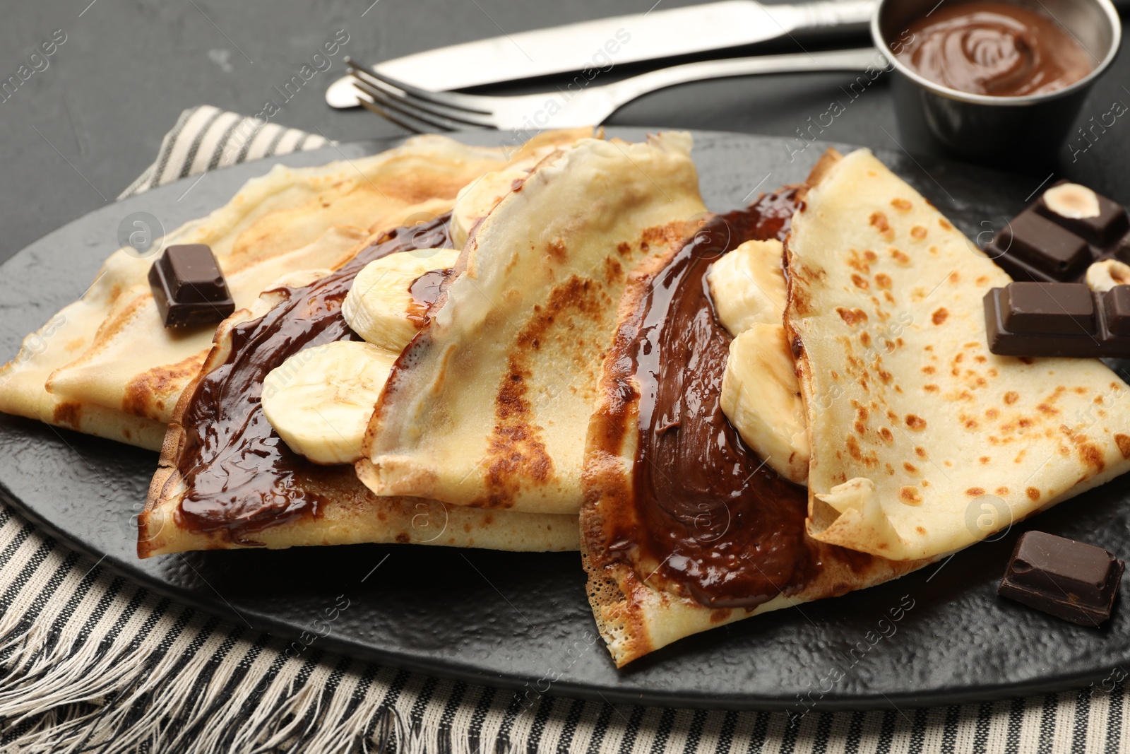 Photo of Tasty crepes with chocolate butter, banana and cutlery on black table, closeup