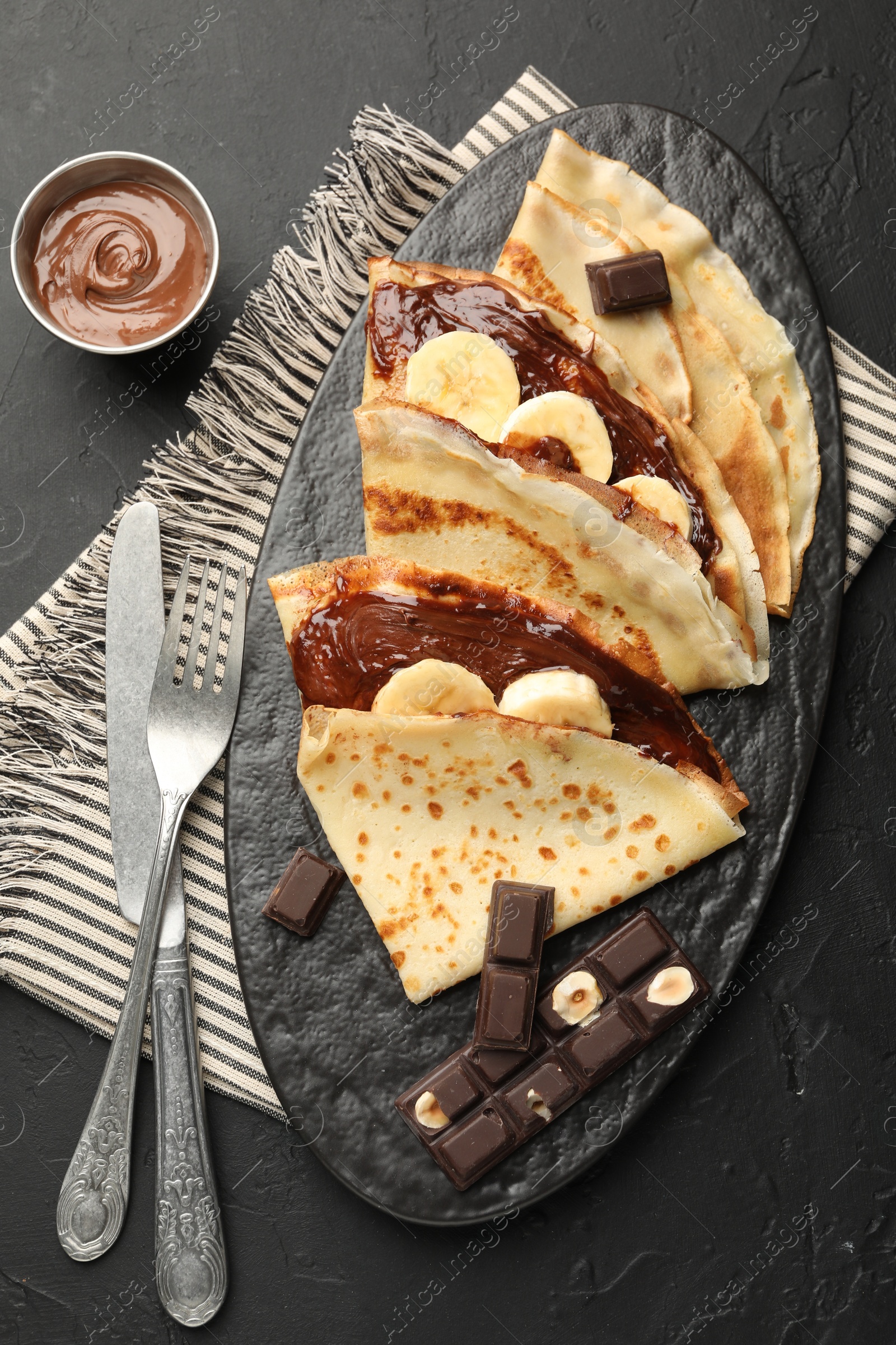 Photo of Tasty crepes with chocolate butter, banana and cutlery on black table, flat lay