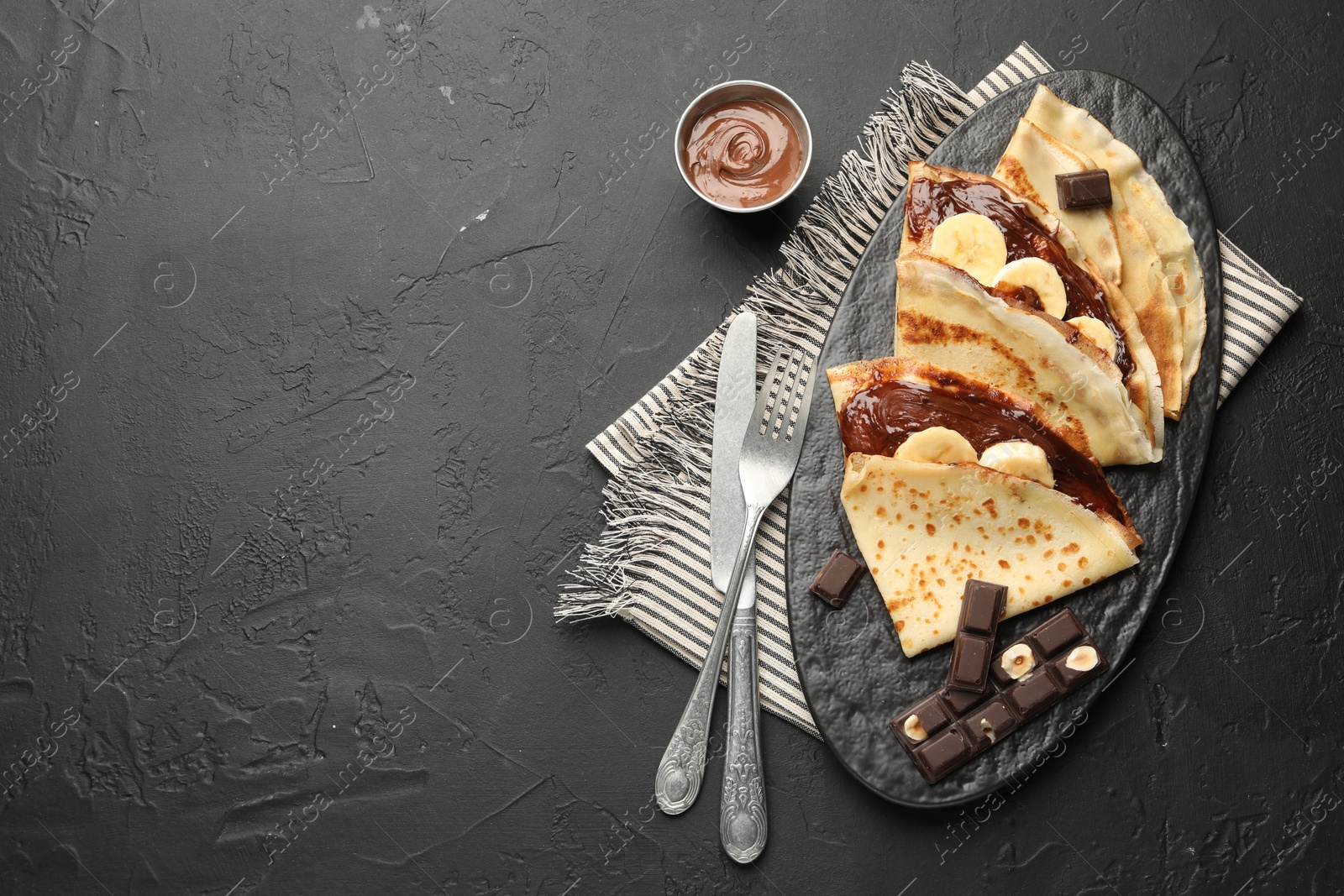 Photo of Tasty crepes with chocolate butter, banana and cutlery on black table, flat lay. Space for text