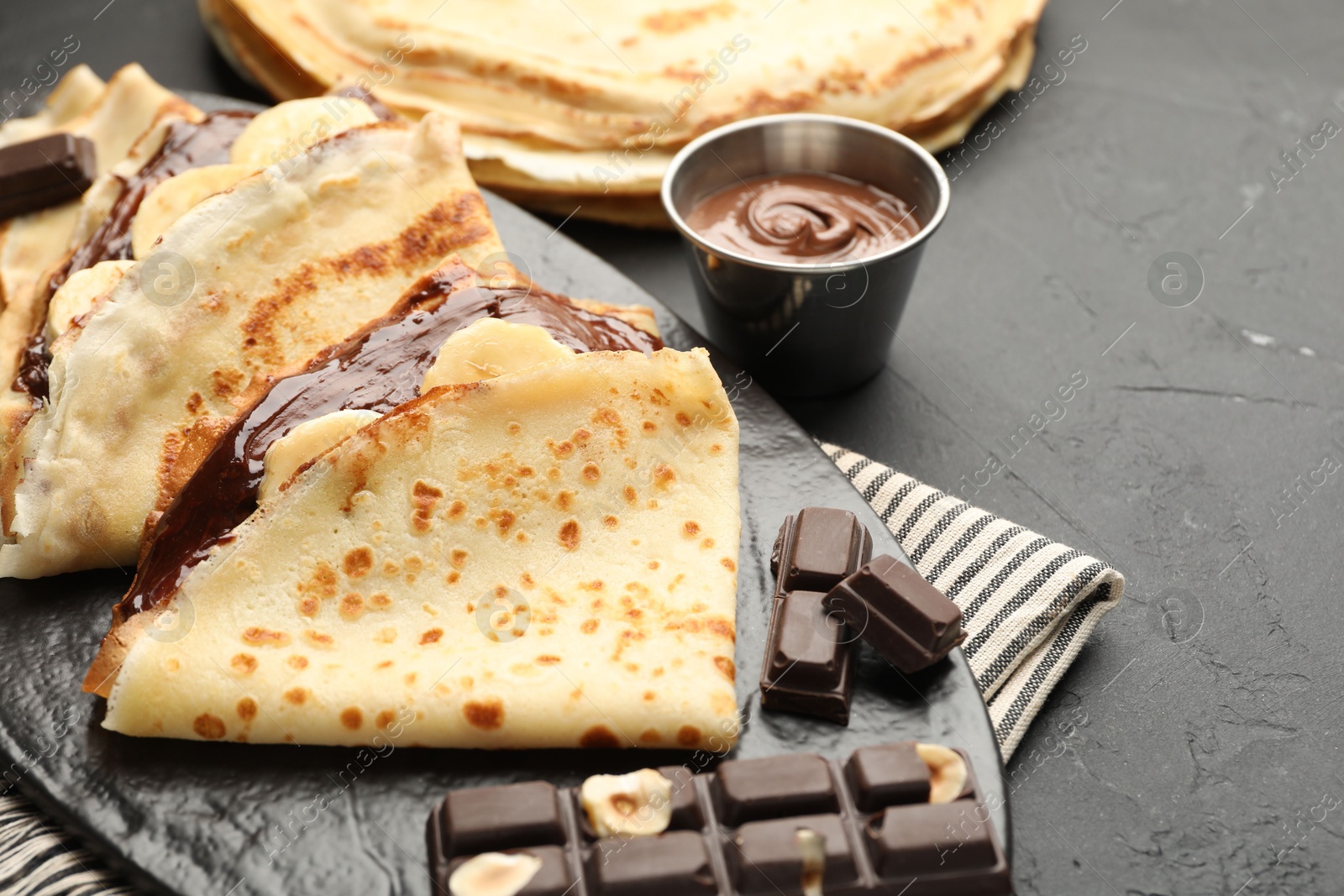 Photo of Tasty crepes with chocolate butter and banana on black table, closeup. Space for text